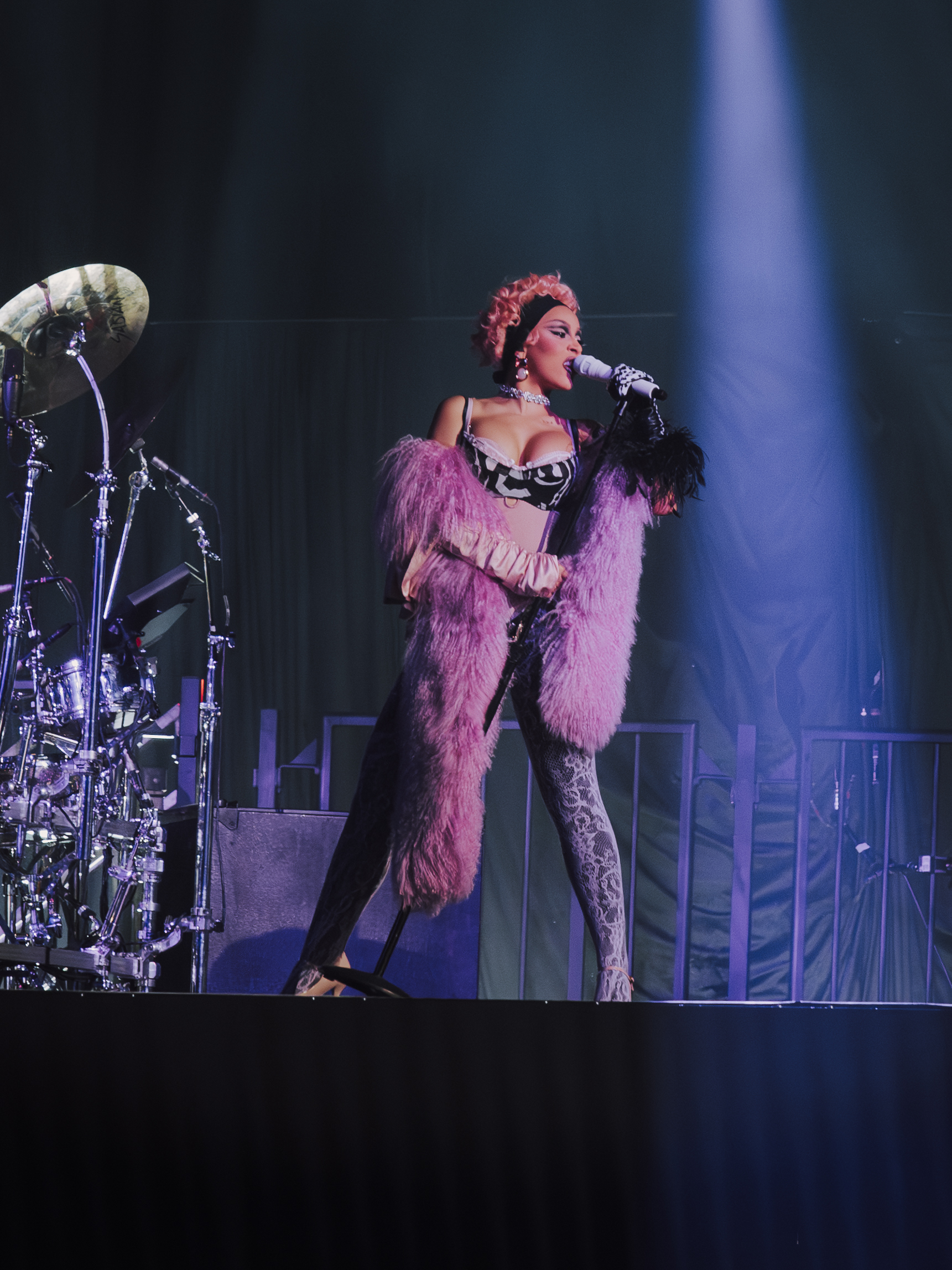 Doja Cat zingt op een podium in een roze bontjas, zwart-witte bustier en strakke broek, terwijl ze een microfoon vasthoudt; naast haar staat een drumstel en een spotlight schijnt van bovenaf op haar.